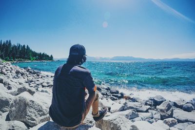 Rear view of man sitting on rocks at beach against sky during sunny day