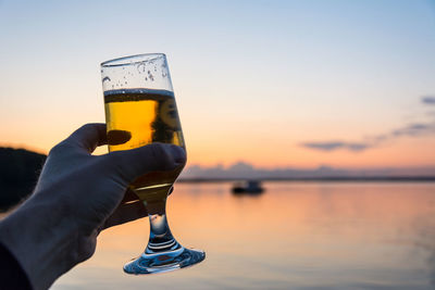 Close-up of hand holding beer glass against sky during sunset