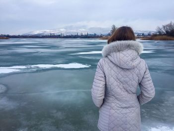 Rear view of woman standing at frozen lake against cloudy sky during winter