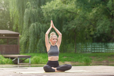 A woman sitting on a wooden platform by a pond in summer, does yoga