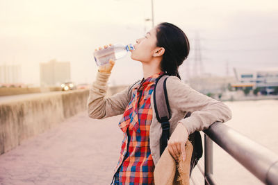 Side view of young woman drinking coffee in city against sky