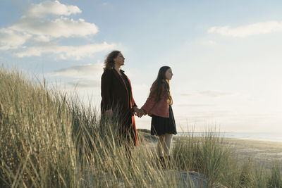 Woman with daughter standing on grass at beach against sky