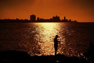 Silhouette man standing by river against sky during sunset in city