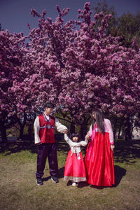 Korean family in national costumes in nature stands next to a cherry blossoming tree.