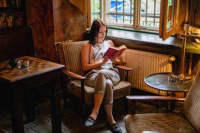 Young woman reading book while sitting on chair by window