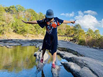 Full length of woman standing standing on rock by water against trees and sky