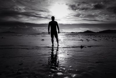 Rear view of silhouette man standing on shore against cloudy sky during sunset