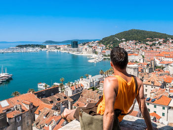 Rear view of man sitting at observation point while looking at sea and city against sky