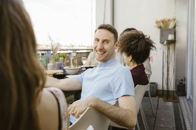 Portrait of happy man sitting on chair with male and female friends while celebrating dinner party in balcony