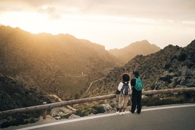 Man and woman looking at mountains while standing near railing on road at sunset