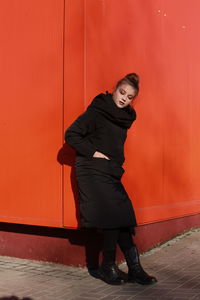 Fashionable teenage girl wearing black warm clothing standing against red wall during sunny day