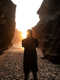 Rear view of man standing by rock formation at beach during sunset