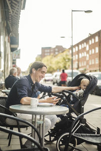 Smiling man listening music through headphones from smart phone while sitting with baby on stroller at sidewalk cafe in