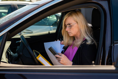 Middle aged business woman in glasses in car working with documents, mobile technology concept