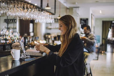 Smiling female entrepreneur with brown hair using laptop while sitting at bar counter