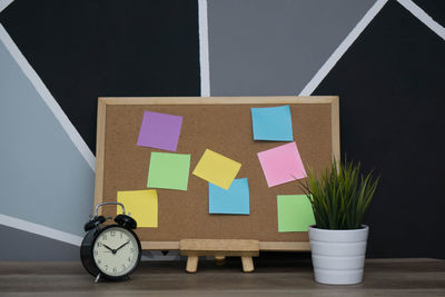 Bulletin board with colorful blank adhesive notes by alarm clock and potted plant on table against wall