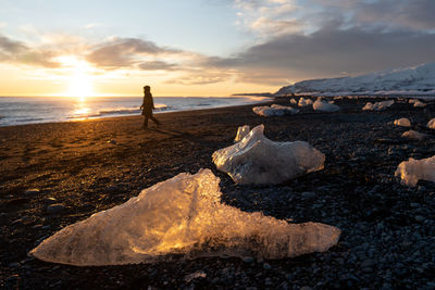 Rear view of man walking on beach against sky during sunset