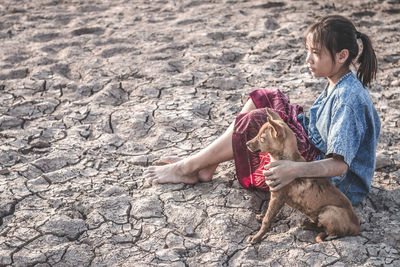 Side view of girl with dog sitting on cracked field