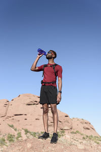 Low angle view of male hiker drinking water while standing on rock formation against clear blue sky