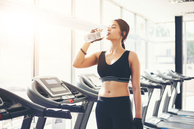 Young woman drinking water while standing on treadmill in gym