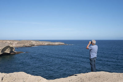 Man looking through binoculars while standing at beach against blue sky