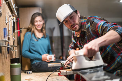 A man waxes his skis while a woman sits on the counter drinking tea in the background