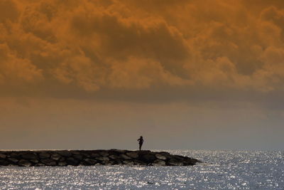 Silhouette person standing on groyne against cloudy sky during sunset