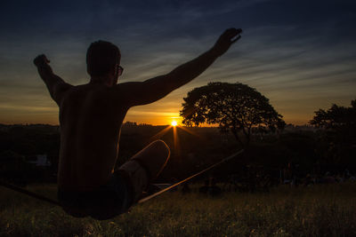 Silhouette man with arms raised sitting on field against sky during sunset