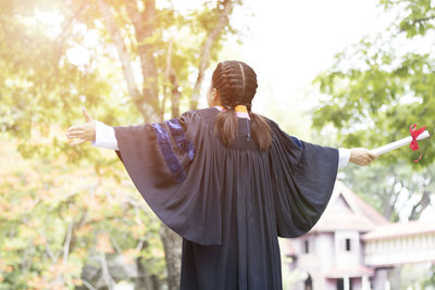 Rear view of woman wearing graduation gown while standing with arms outstretched against trees