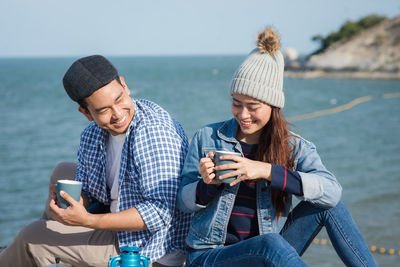 Young couple sitting with mugs while sitting against sea