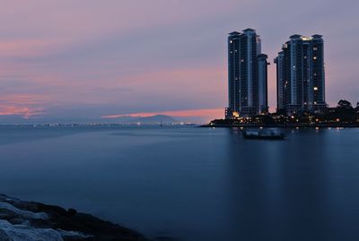 Illuminated city by sea against sky at sunset