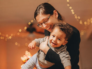 Happy mother carrying cute son against illuminated christmas lights at home