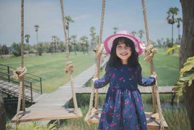 Portrait of smiling girl wearing hat standing by swing against sky