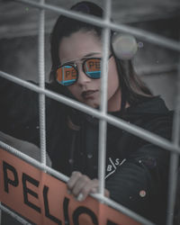 Portrait of young woman wearing sunglasses seen through fence