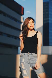 Young woman wearing torn jeans looking away standing against building on terrace 