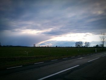 Road passing through field against cloudy sky