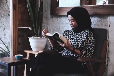 Young woman reading book sitting on chair at home