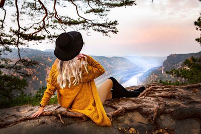 Woman with hand in hair sitting on rock against sky