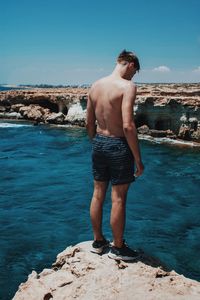 Rear view of shirtless man standing on rock by sea against sky