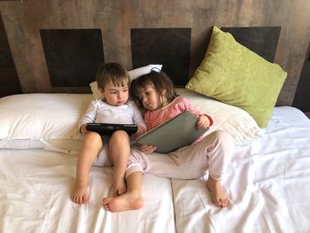 Full length of boy and girl using digital tablet while lying on bed at home