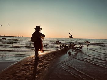 Rear view of woman feeding birds beach against sky during sunset