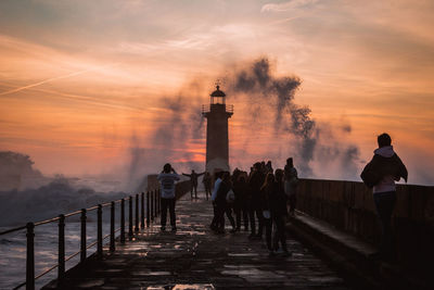 People by lighthouse amidst sea against sky during sunset