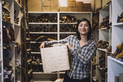 Portrait of smiling female clerk carrying wicker basket while standing amidst shoe racks at store