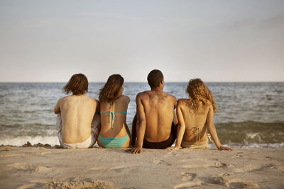 Rear view of friends sitting at beach against sky