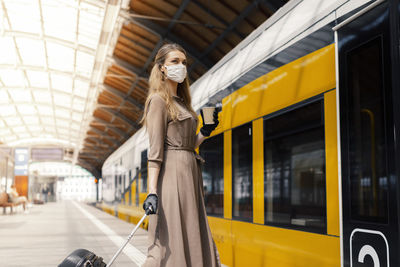 Woman wearing mask holding coffee while standing at railroad station