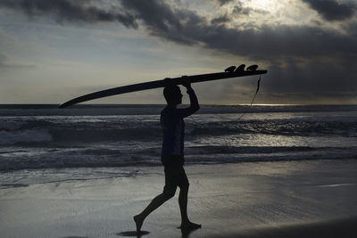 Silhouette man carrying surfboard on head at beach during sunset