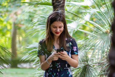 Young woman using phone while standing on palm tree