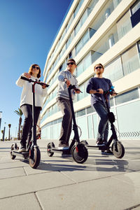 Low angle view of friends using push scooters in city on sunny day