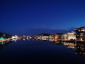 Illuminated buildings by river against blue sky at night