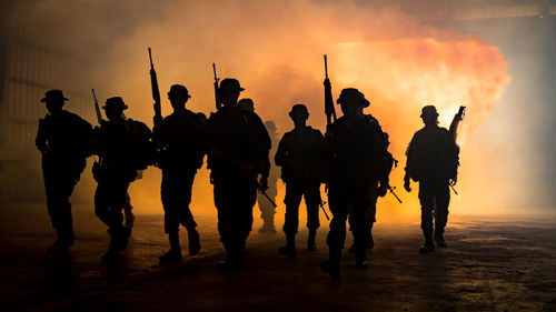 Silhouette people with weapons walking against smoke during sunset